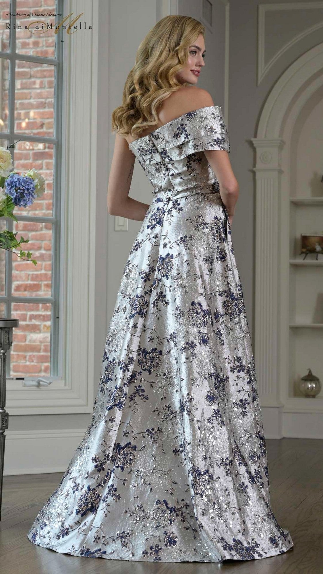 A woman stands indoors wearing the Rina Di Montella RD3425 long formal dress—brocade fabric, off-shoulder neckline, and silver floral pattern—with her back turned to the camera.